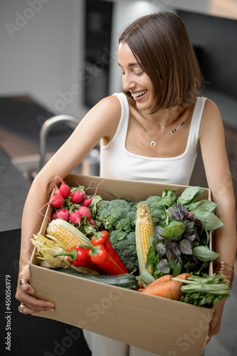 Young woman with fresh vegetables packed in cardboard box at kitchen. Delivering and buying food online, healthy eating and organic food concept
