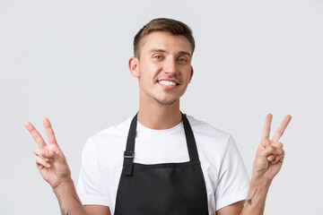 Wall Mural - Cafe and restaurants, coffee shop owners and retail concept. Close-up of friendly cheerful barista, waiter in black apron, showing peace sign and smiling broadly, welcome guests, white background