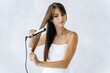 Adorable young woman straightening her hair with a flat iron and looking aside with calm emotions. White wall at the background. Woman hair concept