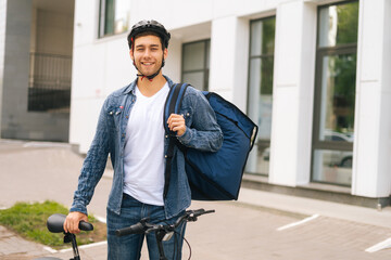 Portrait of cheerful handsome young delivery man in protective helmet posing standing near bicycle in city street, looking at camera. Courier male with thermo backpack delivery food to client