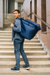 © dikushin - Vertical portrait of cheerful handsome young male of food delivery service with large thermo backpack standing posing in office building stairs, selective focus, looking at camera.