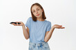 © Cookie Studio - I dont know, not even care. Young woman shrugging while holding mobile phone, looking reluctant and careless at camera, nothing to say, standing over white background