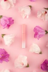 Lip gloss in a pink-to-white gradient case with white and purple hibiscus flowers on a pink background. Flat lay trending scene with buds. Frame made of flowers. Decorative cosmetics liquid lipstick