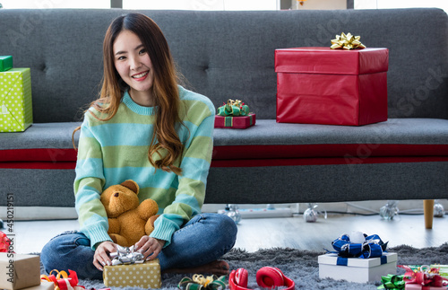 Beautiful young Asian girl on green sweatshirt, smile and sit on floor near sofa in living room with ribbon, gift box, bear doll, red earphone prepared to decorate funny holiday party in winter