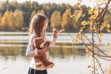 Pretty Little Girl In School Uniform With Cute Teddy Bear Holds Dry Leaf On Riverbank In Sunny Forest On Autumn Day Side View
