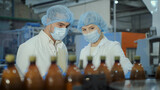 Fototapeta Desenie - Two specialists, a young man and a middle-aged woman, in white coats, disposable hats and medical masks, are discussing production issues against the backdrop of plastic bottles.