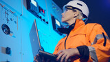 Fototapeta Desenie - Female technician in orange jacket and helmet holds laptop looking at cabinets with switch gears in control office premise close low angle shot