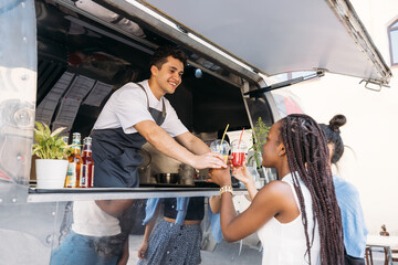 Two customers receiving drinks from food truck owner. Cheerful salesman in apron looking at clients.