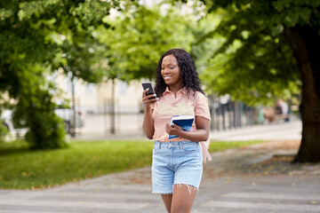 Wall Mural - technology, education and people concept - happy smiling african american student girl with smartphone and book in city