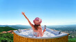 © Goffkein - Young carefree girl traveler relaxing with a glass of wine at hot tube during enjoying happy traveling moment vacation life against the background of green big mountains