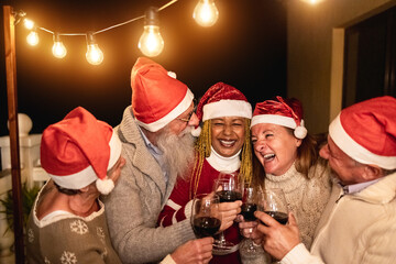 Wall Mural - Diverse senior people having fun cheering with wine during Christmas eve wearing Santa Clause hats - Soft focus on center man face
