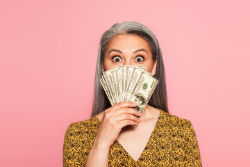 amazed middle aged woman covering face with dollar banknotes isolated on pink