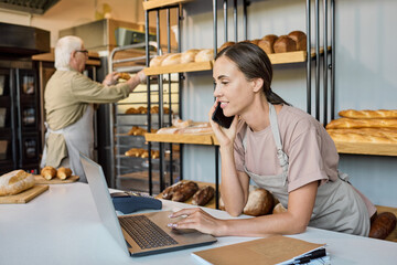 Busy female in apron using laptop and talking to clients on the phone by workplace