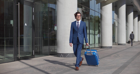 Young stylish businessman walking with suitcase near airport building