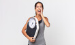 © kues1 - young hispanic woman feeling happy,giving a big shout out with hands next to mouth and holding a weight scale. diet concept
