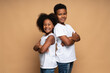Waist up portrait view of the lovely multiracial brother and sister standing back to back to each other and smiling while posing at the studio over the beige wall