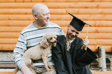 Senior Elderly Man Grandfather Holds Poodle Dog In His Arms And Hugs His Grandson Boy Who Graduate Of Elementary School In Cape And Graduate Cap On Porch Of Rustic Wooden House, Concept Of Graduates