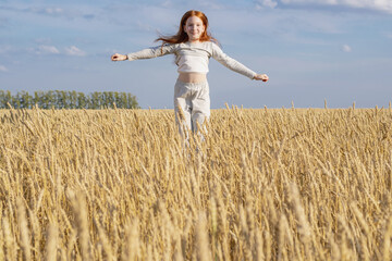 a little young girl with red hair flies over a golden wheat field against the blue sky.