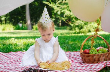 Adorable caucasian blonde baby girl celebrating first birthday outdoor at picnic in summer park ,wearing festive hat,with balloons,fruits,croissants.Kid having meal,festive lunch, party for infant