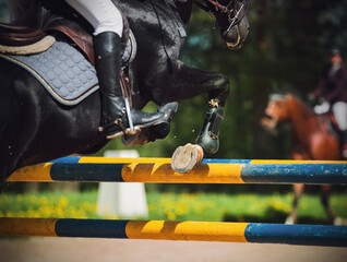 A black horse with a rider in the saddle jumps over a high yellow-blue barrier at a show jumping competition on a sunny summer day. Horse riding. Equestrian sports.