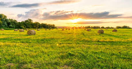 Scenic view at sunset or sunrise in green shiny field with hay stacks, bright cloudy sky, golden sun rays, summer valley landscape