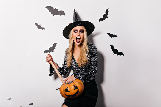 Stylish witch in big hat holding pumpkin and screaming. Adorable blonde vampire posing with aspen stake.