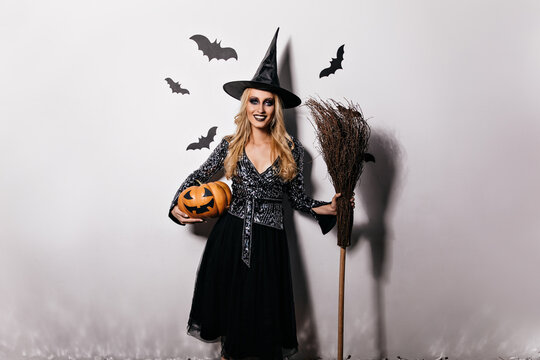 Romantic blonde girl posing with pumpkin and bats. Indoor shot of smiling young woman enjoying halloween carnival.