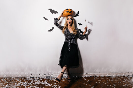Full-length portrait of emotional woman in black dress posing after carnival. Surprised blonde girl holding halloween pumpkin.
