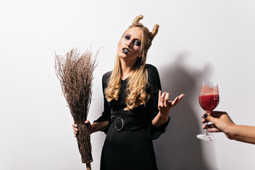 Wall Mural - Bored blonde witch looking at goblet with blood. Fair-haired vampire posing in studio with broom.
