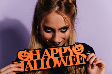 Wall Mural - Sinister fair-haired girl looking to camera with smile. Pleased young woman with scary dark makeup posing on purple background in halloween.