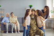Happy veteran soldier comes back from the military and reunites with family. Wife and two children who missed dad kissing him on cheeks standing on blurred copy space home background with grandparents
