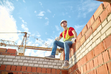 Poster - Cute tranquil builder in a hardhat seated on a brick structure