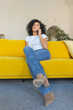 © kinomaster - Portrait of a young African American woman talking for mobile phone. Brunette with curly hair sitting on yellow sofa in a bright home room.