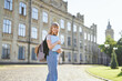 Adorable happy young blonde university or high school girl student with backpack and laptop wearing jeans standing in university campus. Educational or studying concept. High quality image