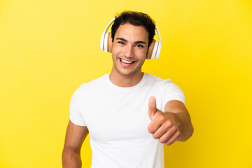 Caucasian handsome man over isolated yellow background listening music and with thumb up