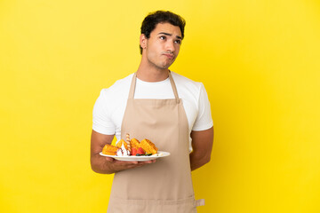 Sticker - Restaurant waiter holding waffles over isolated yellow background and looking up