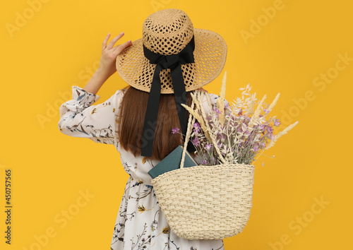 Woman holding beach bag with beautiful bouquet of wildflowers and book on yellow background, back view