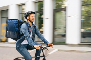 Wall Mural - Side view of happy handsome young delivery man with thermo backpack riding bicycle in city street on blurred background of office building. Courier male in protective helmet delivery food to client