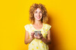 © Alex - curly young woman smiling holds a phone on a yellow background.