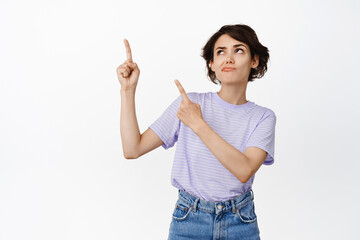 Wall Mural - Unsure, indecisive brunette woman frowning pensive, pointing fingers and looking at upper left corner, advertisement up, hesitating, being puzzled, white background