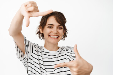 Wall Mural - Happy smiling woman looking through hand frames, taking picture, standing against white background