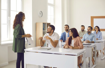 College teacher having class with group of interested adult male and female business course students. People sitting at tables in classroom, discussing topic of success, asking or answering questions