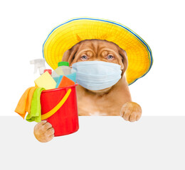 Poster - Puppy wearing panama and medical protective face mask and red hat  looks from behind empty white banner and holds bucket with  washing fluids and rags in paws. isolated on white background