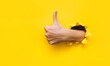 The female hand shows a thumb up gesture (like). Torn hole in yellow paper. The concept of positive attitude, approval and praise. Copy space.