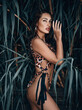 Young sexy brunette woman savage in leopard print pattern bikini standing sideways at, hiding behind tropical tree leaves, holding hand at head and looking at camera. Summer vibes