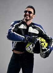 Portrait of happy laughing bearded man biker motocross race winner in sunglasses, motorcycle gear leather jacket, jeans and gloves stands holding his helmet in hands over light background