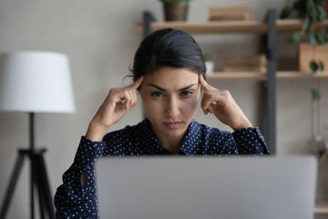 Wall Mural - Pensive focused millennial Indian woman look at laptop screen think ponder of problem solution. Concentrated young biracial female employee work on computer make decision or plan at workplace.