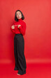 Vertical shot of Asian woman stands indecisive and thoughtful makes unsure gesture keeps index fingers opposite each other wears poloneck black bell bottomed trousers isolated over red background