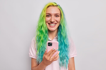 Happy teenage girl glad to download application on smartphone smiles happily has long dyed hair piercing in nose dresses casually isolated over grey background. Youth and technology concept.