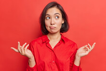 Clueless Hesitant Beautiful Asian Woman With Short Dark Hair Spreads Palms Sideways Shrugs Shoulders Has Indecisive Expression Dressed In Shirt Stands Against Vivid Red Background. Who Knows.